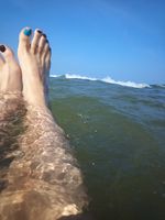 Two white feet floating in large body of water with a foaming wave. The sky behind is blue. The toenails are painted blue and brown.
