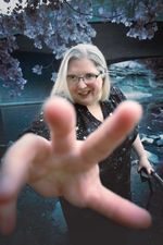A white person with blonde hair, glasses and a cane, reaches their fingers towards the camera to draw you into their world. Beyond them lies a silvery tree and a creek.