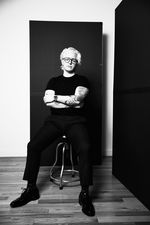 Full body photo of Leone "Erikx" DiSantis sitting on a stool with crossed arms. They have light skin, sideswept short white hair, aviator-style glasses and tattoos on their arms. They wear an all-black look of t-shirt, pants, watch, and oxfot-style shoes.