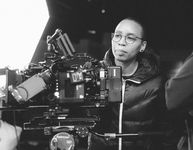 Black & white photo of Melinda James staring at a large videocamera.  Melinda is a Black and Thai person with round glasses and a focused expression.