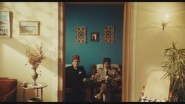 Two Arab women wearing suits and fez hats in different colours sit with direct eye contact with the camera as they recline on lounge chairs in a vibrant room with cultural artwork and decorations. The camera is positioned in the doorway of a neutral coloured room, creating an artistic frame for the women.