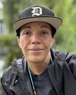 Raven looks directly at the camera with a slight smile and warm eyes. They have brown eyes and freckles. They have short black hair and are wearing a black ballcap with a stylized D. They also wear a back raincoat and a thin necklace with turqouise, black, red, yellow and white beads.