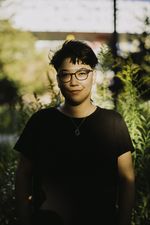 Kay looks directly at the camera with a confident yet sly smile. They have light skin, short dark hair, and dark brown eyes. They are wearing dark grey glasses, a black shirt, and a necklace. Behind htem are some blurry young trees.
