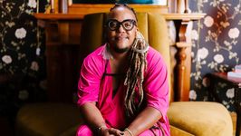 Syrus sits on a plush yellow chair with a welcoming expression. Syrus is a Black trans man with thick-rimmed glasses, a beard, and dreadlocks cascading over his shoulder. He wear a hoop earing, a feather earring, and a hot pink fashionable onesie.