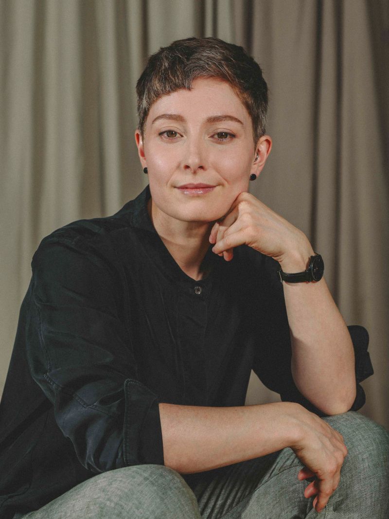 Cailleah has light skin and short, light-brown hair. They're wearing a dark shirt and watch, and they're smiling with their chin resting on their hand.