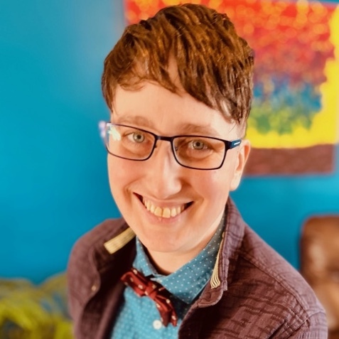 Picture of Danielle Peers, who has short brown hair, black rectangular glasses, and is wearing a brown jacket over a blue button down shirt and red polka dot bow tie. They are smiling at the camera in front of a colourful, out of focus backdrop.