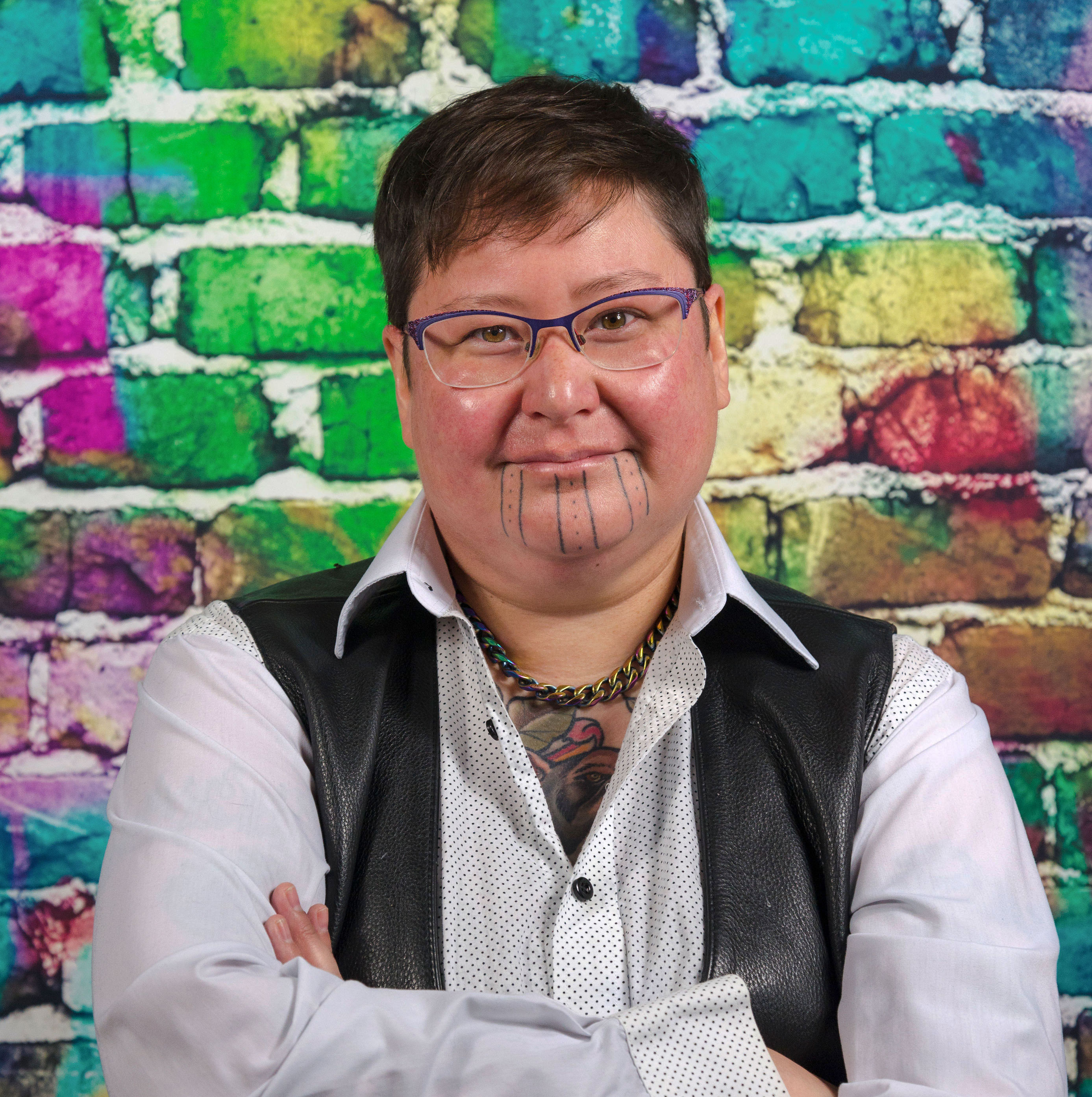 TJ has light skin, short hair, glasses, and three vertical lines tattooed on his chin. He stands in front of a rainbow brick wall wearing a light button-down shirt and a leather vest.