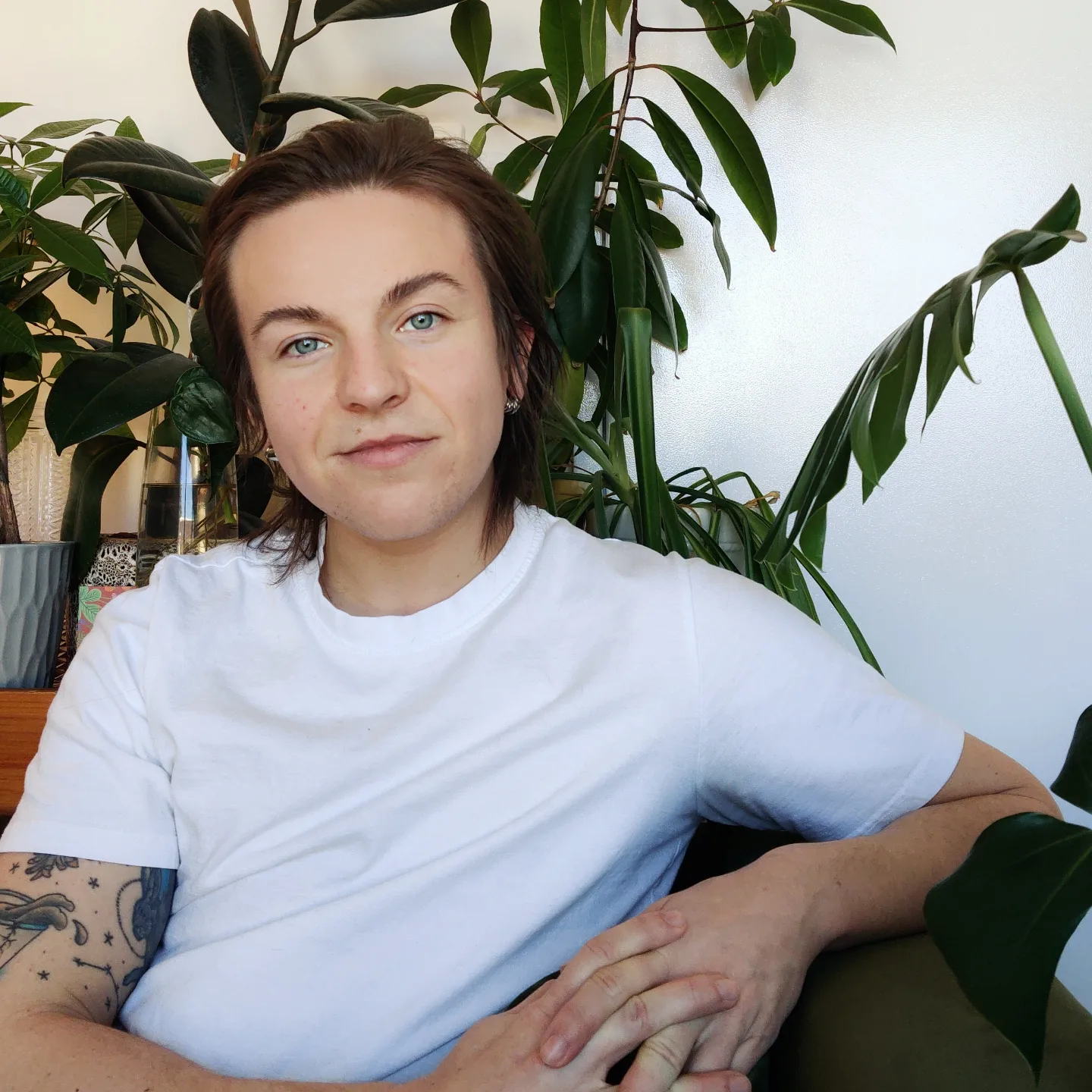 Kit is a white masculine person with shoulder-length brown hair. They are wearing a white t-shirt and are sitting with their hands folded in front of them, looking into the camera.