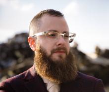 Outdoor photo of white trans man with glasses and prodigious brown beard