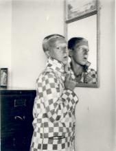 Claude Cahun dressed in a large checkered jacket in front of a mirror looking toward the camera; black and white image.